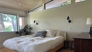 A bedroom with a large bed, a nightstand, a lamp, and a window with a view of trees. - Centrally Located Bright Family Home with Peaceful Natural Views and Central A/C (Burlington)