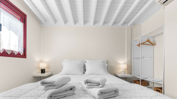 Een net opgemaakt bed met witte lakens en een bedekking met patroon, een nachtkastje met een lamp, en een kledingkast met hangruimte.