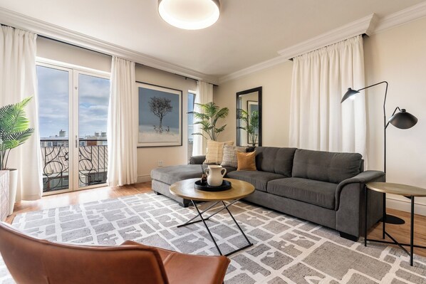 A modern living room with a grey sofa, a round coffee table, and a large window with curtains. - Stylish Green Point Apartment | Rooftop Pool (Cape Town)