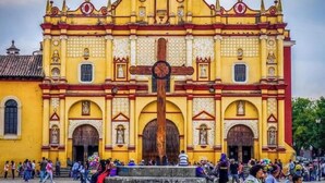 A historic yellow building with a large wooden cross in front, surrounded by tourists.