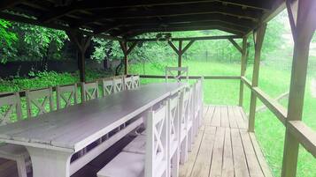 A covered picnic area with a long table and benches, surrounded by a wooden deck and a grassy lawn.