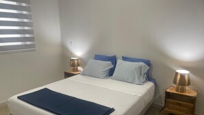 Een net opgemaakt bed met een blauwe deken, twee bedlampjes en een raam met jaloezieën.