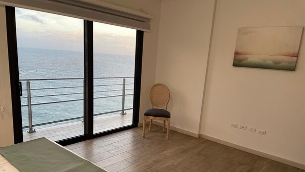 Een kamer met een grote schuifpui naar de zee, een houten vloer, een enkele stoel en een schilderij aan de muur.
