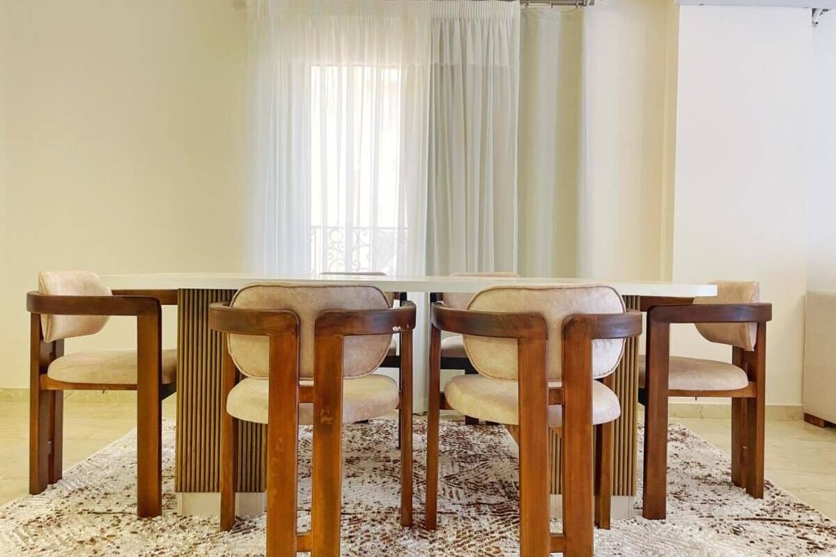 A dining area with a rectangular table and six chairs, a patterned rug, and a window with sheer curtains.