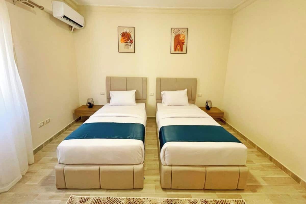 A twin room with two beds, a small table, a nightstand, an air conditioning unit, and two wall art pieces.
