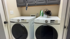 Dos lavadoras Samsung de carga frontal en un cuarto de lavado con un cerrojo en la puerta, un planchador verde y un adorno en la encimera.