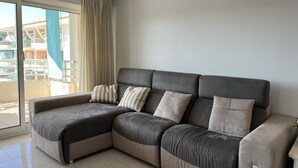 A modern living room with a sectional sofa, a sliding glass door leading to a balcony, and a view of a building with balconies.