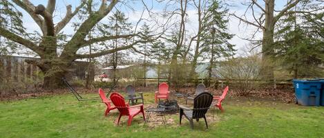 Un jardin avec un foyer, des fauteuils Adirondack rouges et noirs, et une poubelle bleue.