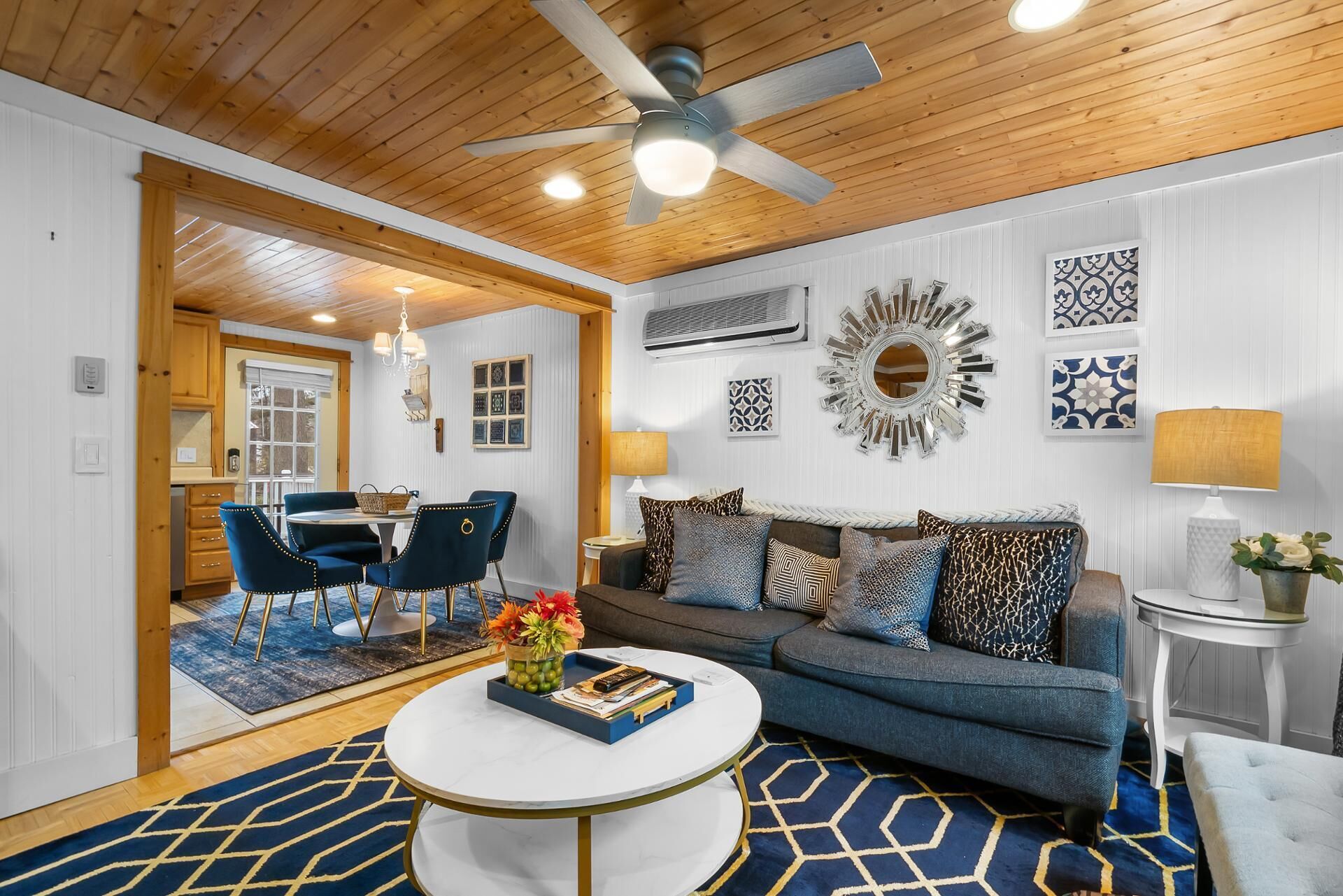 Un salon bien éclairé, doté d’un ventilateur de plafond, un coin repas avec des chaises bleues, un canapé bleu avec des coussins à motifs et une table basse ronde.
