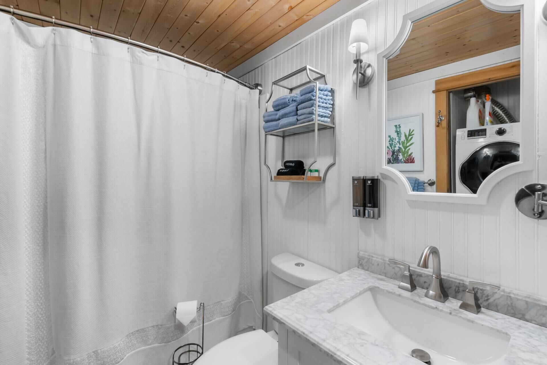 Een moderne badkamer met douchegordijn, toilet, wastafel met marmeren blad, handdoekrek met opgevouwen handdoeken, toiletpapierhouder en wandlamp.