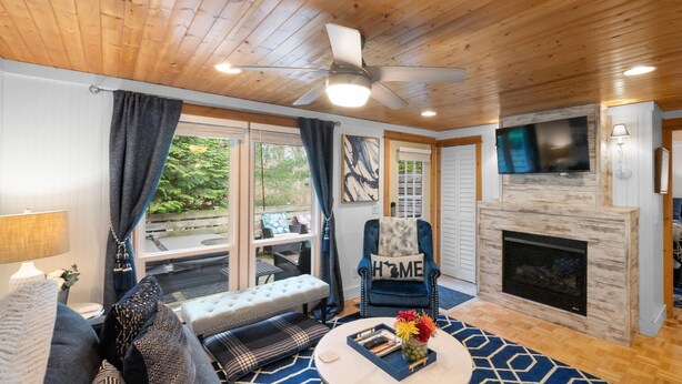 A cozy living room with a fireplace, a ceiling fan, and a large window with curtains. - "cozy Cottages" Blue Cottage-hot Tub/town! (Saugatuck)