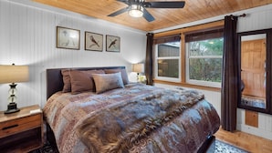 Une chambre avec un plafond en bois, un lit recouvert d’une couverture à motifs, deux lampes de chevet, une fenêtre avec des rideaux et trois tableaux encadrés accrochés au mur.