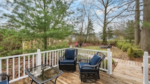 Une terrasse en bois, agrémentée de mobilier d’extérieur, notamment deux chaises et une table, entourée d’arbres et d’une clôture blanche.