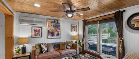 A cozy living room with a wooden ceiling, a ceiling fan, a brown leather sofa, a coffee table, and a large window with a view of greenery.