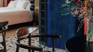 A modern hotel room with a bed, a desk, and a chair. There is a large window with curtains, a decorative vase with flowers, and a blue accent wall. - Apartament przy MCK (Katowice)