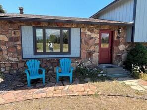 A stone house with a red door, two blue Adirondack chairs, and a window reflecting a person.