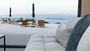 Un área de estar moderna al aire libre con sofá, sillones reclinables y sombrillas, con vista a un cuerpo de agua y montañas distantes.