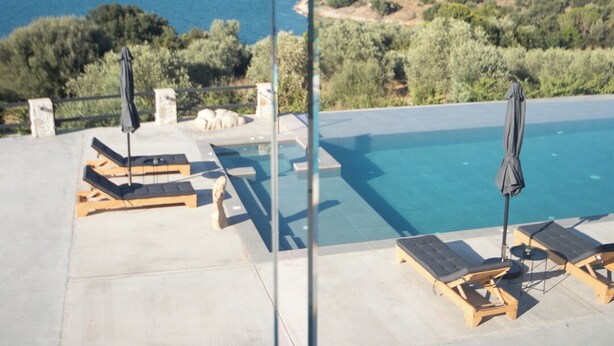 Zona de piscina con sillones de descanso y sombrillas, con vista a un cuerpo de agua y follaje.