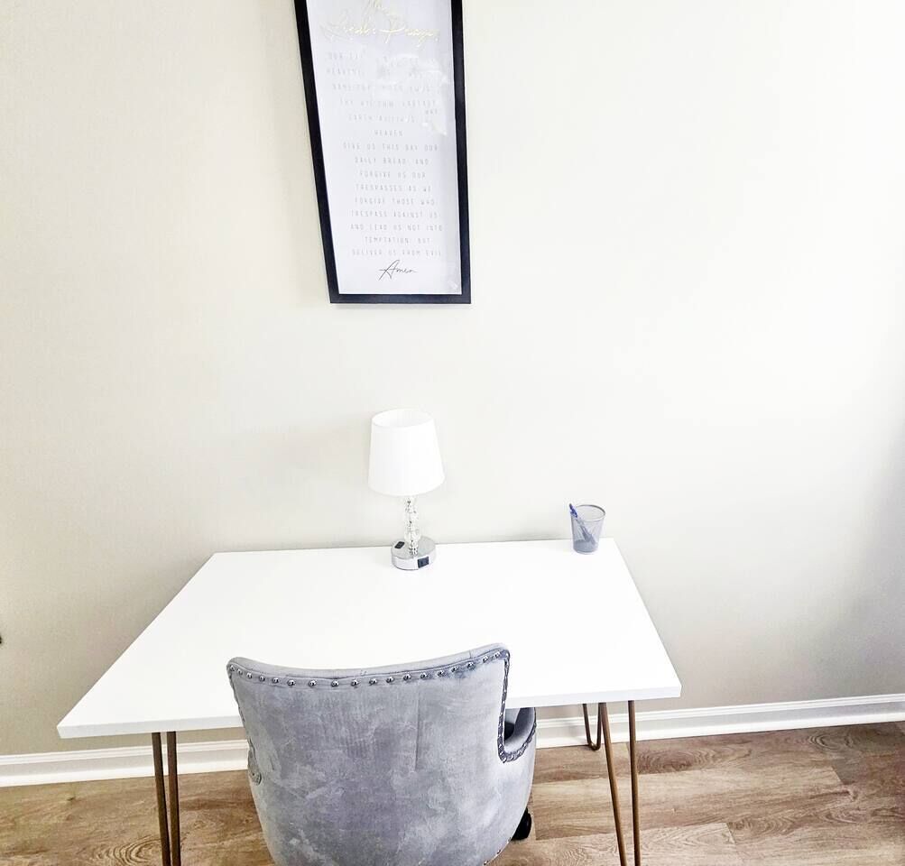 Habitación minimalista con una mesa blanca, una silla gris, una lámpara blanca y un texto enmarcado en la pared.