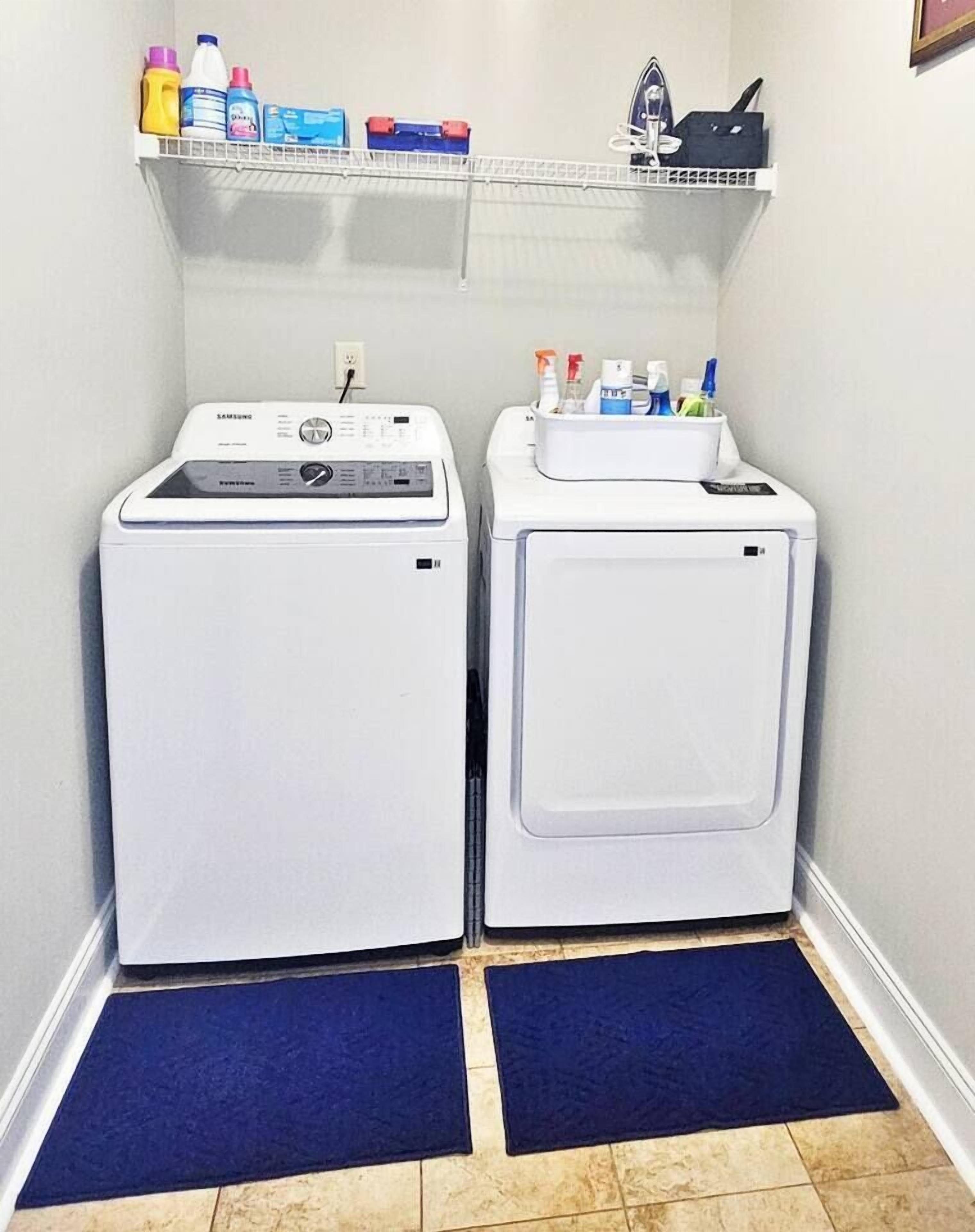 Un área de lavandería con dos lavadoras blancas, un estante con productos de limpieza y un tapete azul frente a cada máquina.