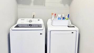 Un área de lavandería con dos lavadoras blancas, un estante con productos de limpieza y un tapete azul frente a cada máquina.