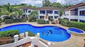 A hotel with a swimming pool surrounded by lush greenery and multiple-story buildings with blue accents. - 2-bedroom condo in beautiful Playa Del Coco.  5 minute walk to beach (Guanacaste Province)