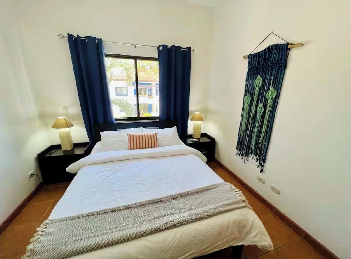 A neatly made bed with a white comforter and a gray throw blanket, two bedside lamps, a window with blue curtains, and a wall-mounted curtain.