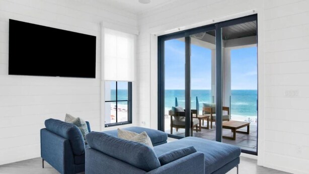 Ein modernes Wohnzimmer mit einem großen Fernseher, einem blauen Sofa und Meerblick durch Glastüren.