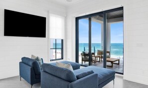 A modern living room with a large TV, a blue sofa, and a view of the ocean through glass doors. - Sea La Vie at Eleanor Beach (Pensacola)