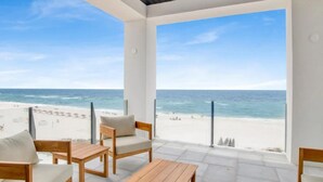 Eine Terrasse am Strand mit Blick auf das Meer und den Strand, ausgestattet mit Holzmöbeln und großen Fenstern.