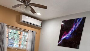 Un dormitorio con cama, ventilador de techo, aire acondicionado, ventana con cortinas y un cuadro colgado en la pared.