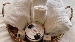 Una cama con ropa blanca y una cesta tejida que contiene una vela, una caja con la etiqueta 'Dove' y toallas dobladas.