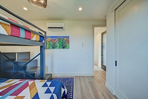 A room with bunk beds, a colorful quilt, a wall-mounted air conditioner, and a door leading to another room.