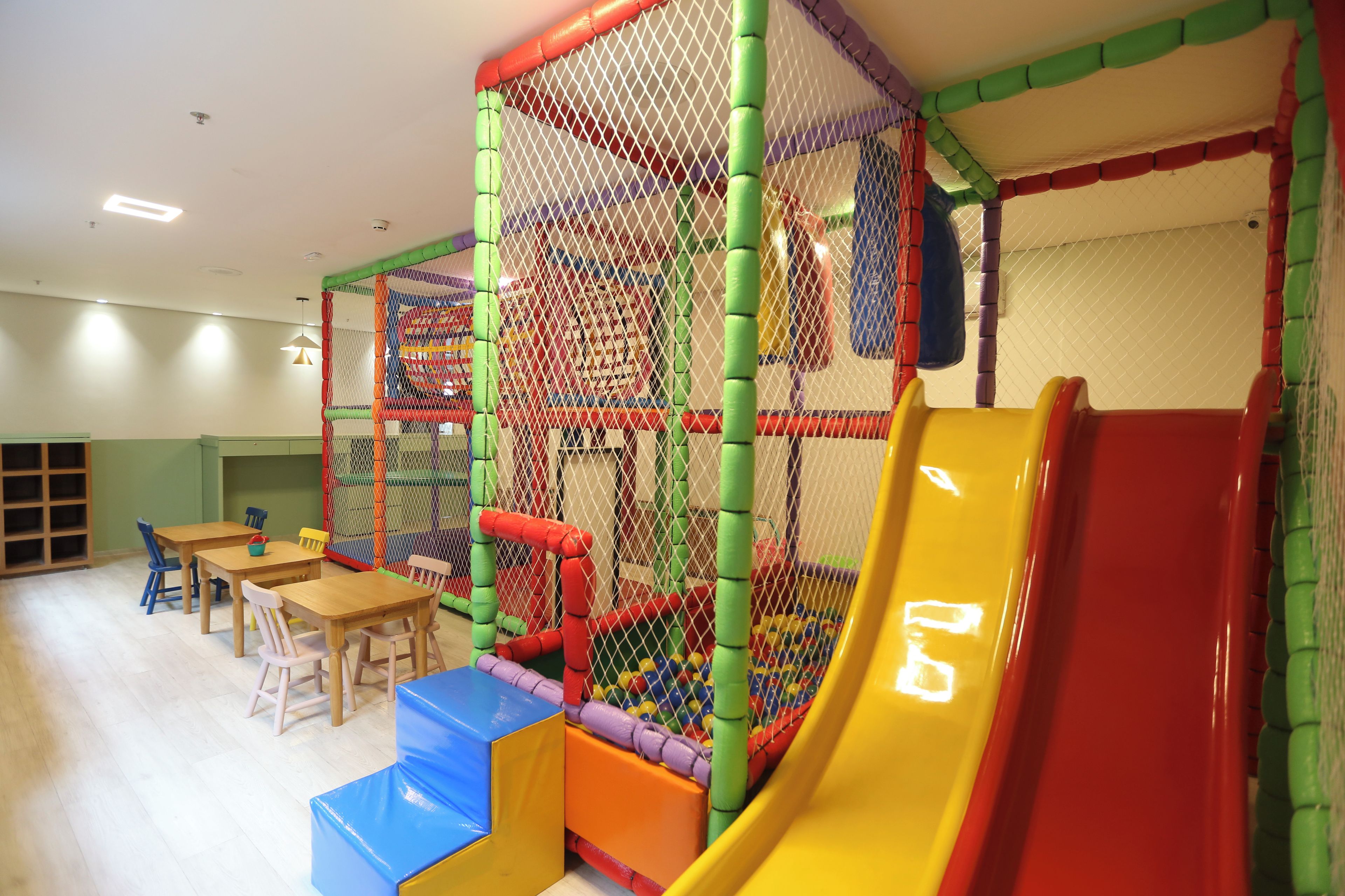 A children's play area with a slide, soft play structures, and tables with chairs.
