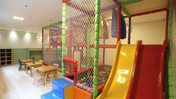 A children's play area with a slide, soft play structures, and tables with chairs.