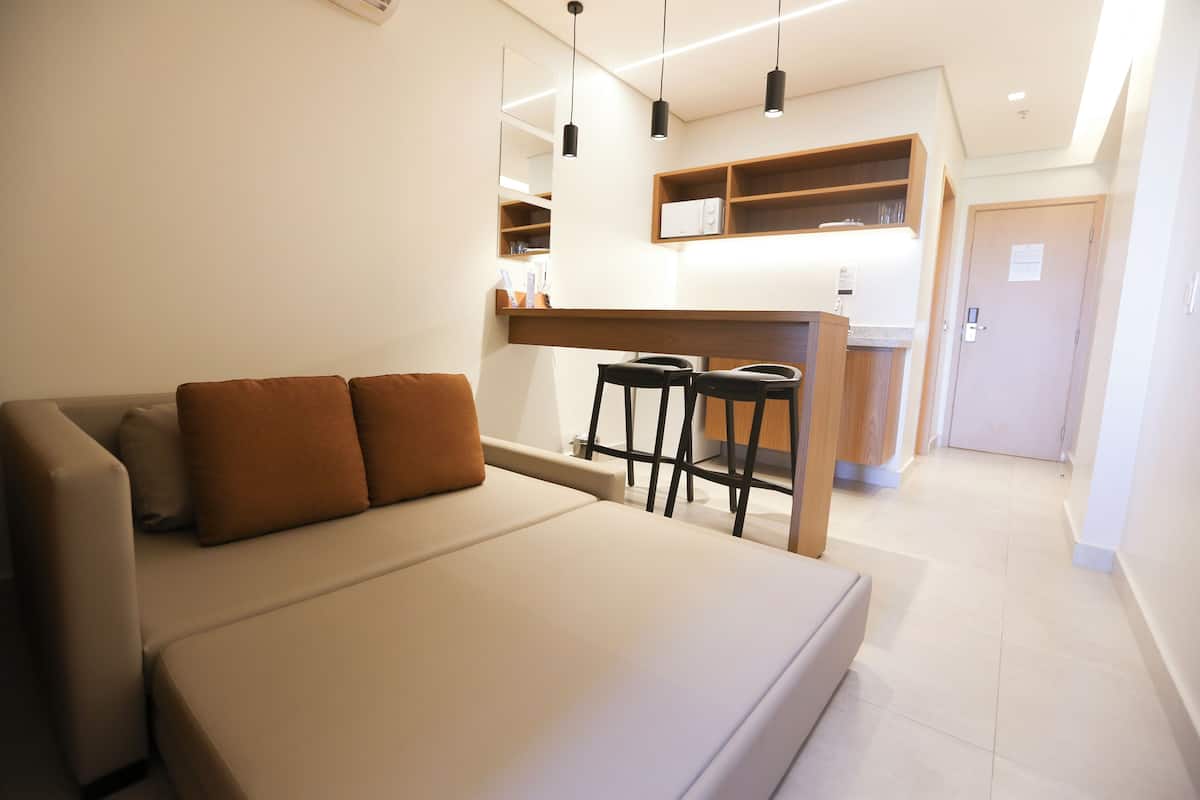 Ein modernes Hotelzimmer mit einer Sitzecke, einer kleinen Bar mit Hockern, einer Küchenzeile und einer Tür zu einem weiteren Raum.