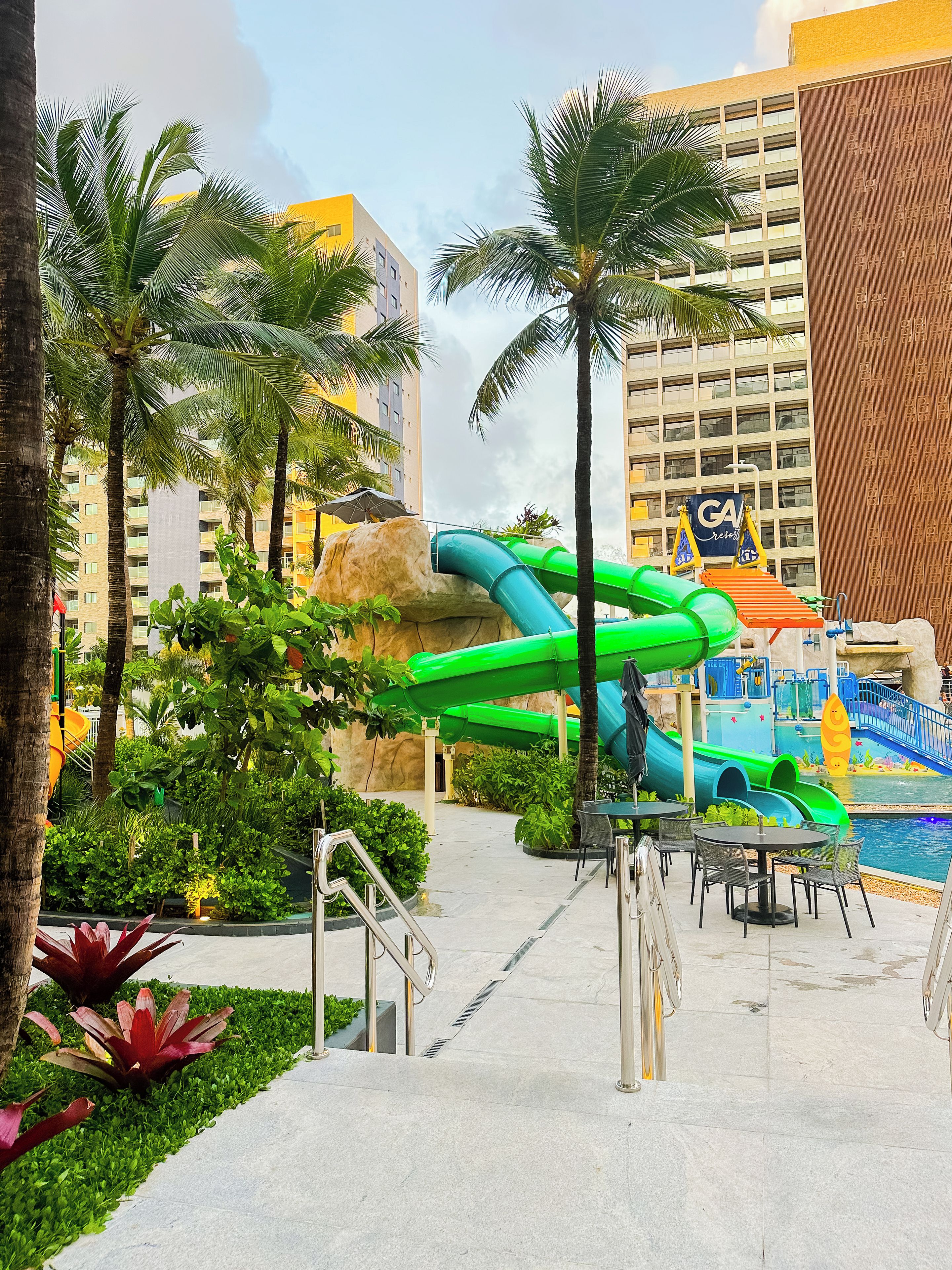 A water park area with slides and palm trees, surrounded by buildings.