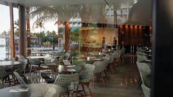 Un restaurante con mesas redondas y sillas de mimbre, amplias ventanas y vistas a palmeras.