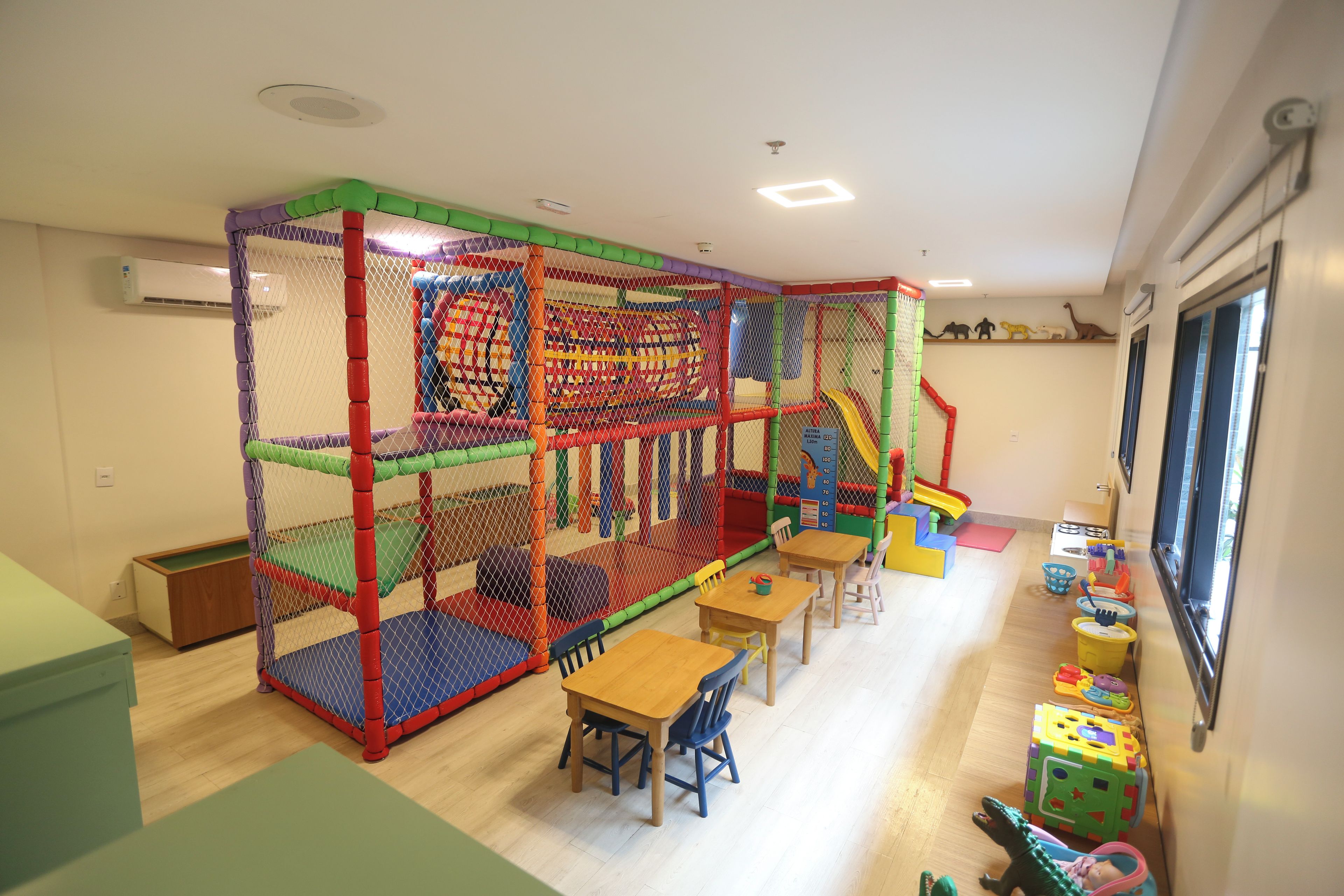 Zona de juegos con cerramiento de red, mesas, sillas y estructuras de juego coloridas.