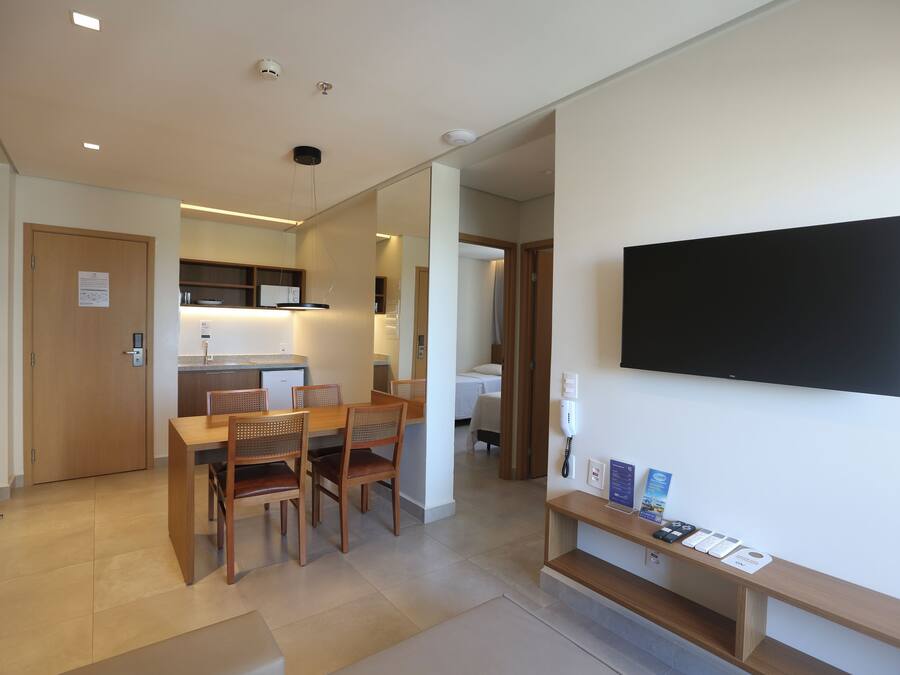 Una habitación de hotel moderna con zona de comedor, un televisor de pantalla plana en la pared y un pequeño estante con folletos informativos.