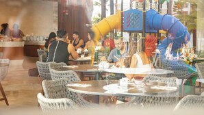 Un ristorante con tavoli rotondi e sedie in vimini, una struttura giochi all'esterno e un bancone con il personale.