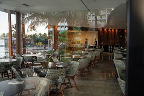 Un ristorante con tavoli rotondi e sedie in vimini, ampie vetrate e vista su palme.