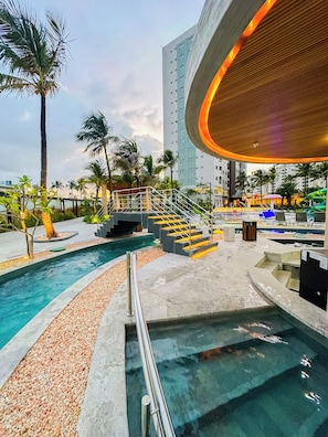 Un área de piscina con agua cristalina, palmeras y un edificio moderno al fondo.