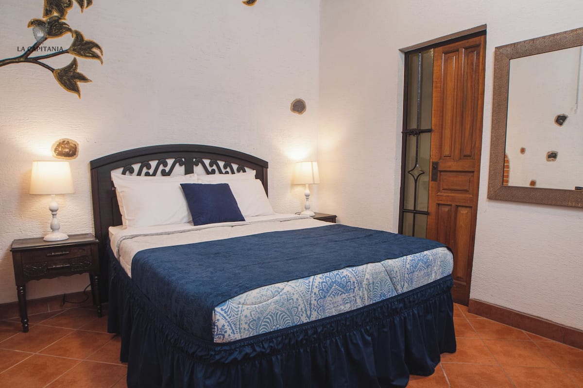 A bedroom with a bed, two bedside tables with lamps, a wooden door, and a wall-mounted mirror.
