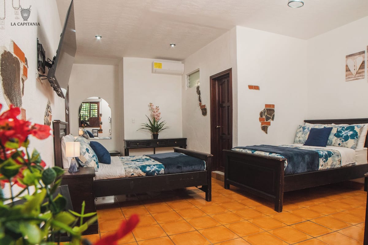 A room with two beds, a desk, a chair, a potted plant, and a wall-mounted TV.
