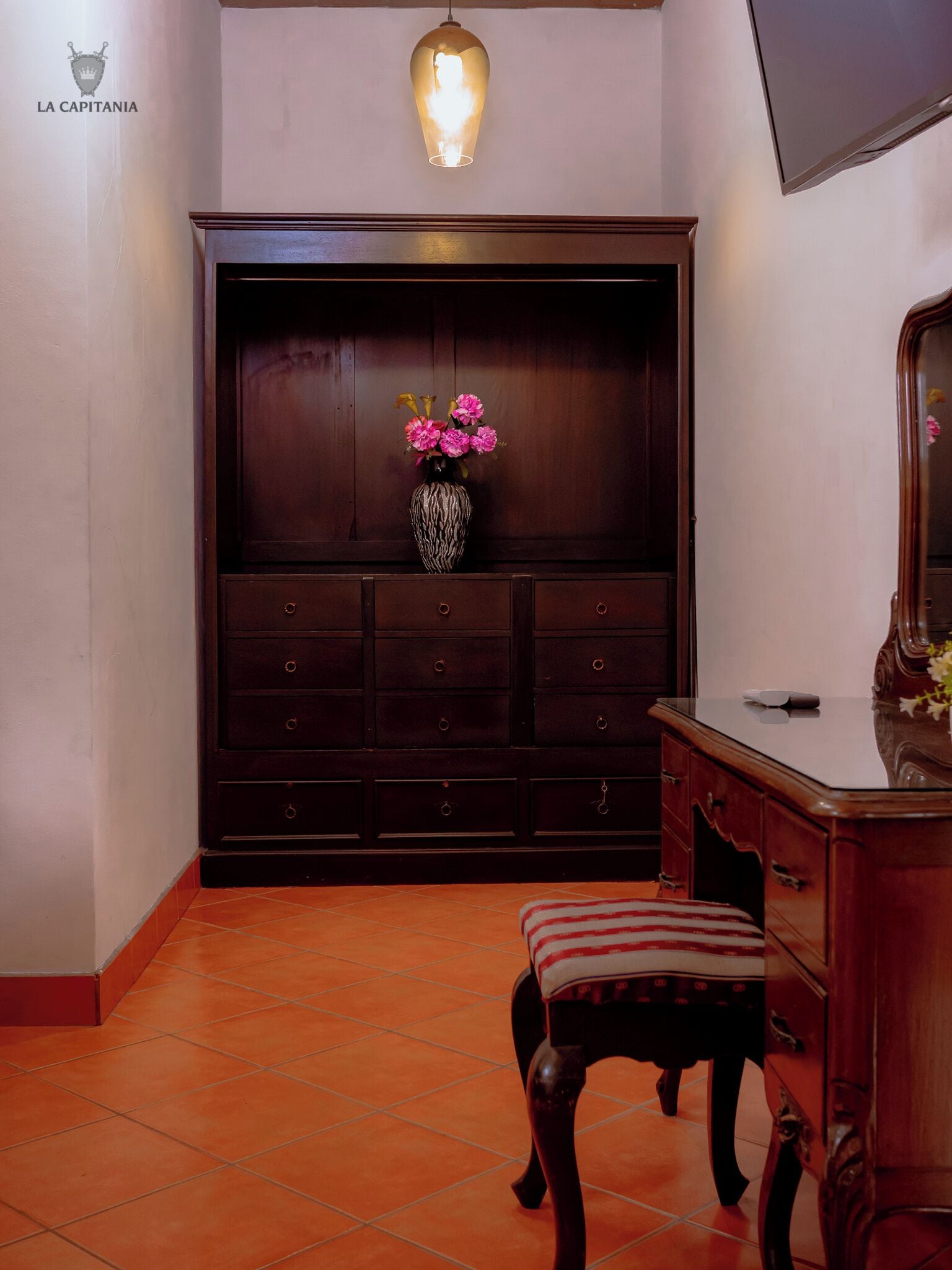 A room with a dark wooden cabinet, a vase with pink flowers, a hanging light fixture, and a tiled floor.