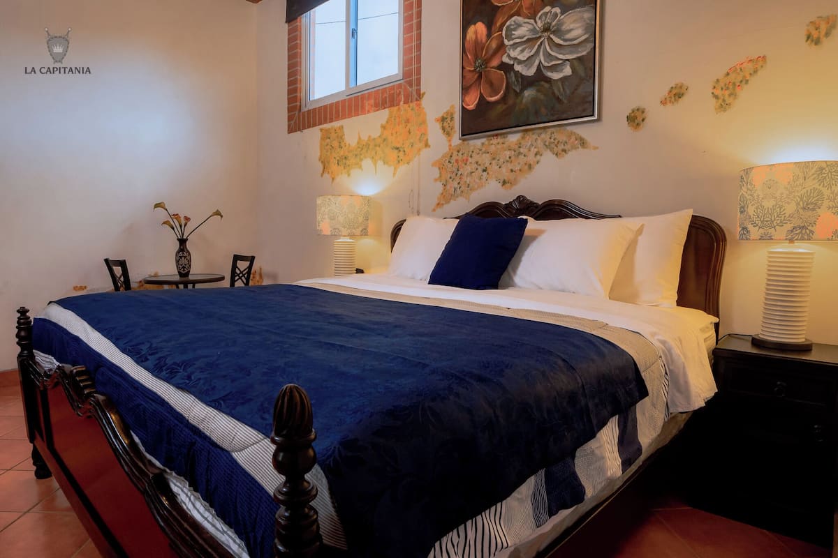 A hotel room with a bed, bedside tables, a nightstand, a window, and a wall with a floral painting.