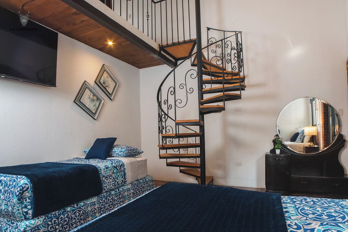 A bedroom with a spiral staircase, two beds with patterned bedding, a large mirror, and a flat-screen TV mounted on the wall.