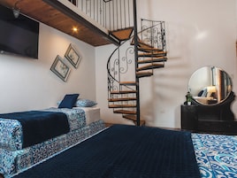 A bedroom with a spiral staircase, two beds with patterned bedding, a large mirror, and a flat-screen TV mounted on the wall.