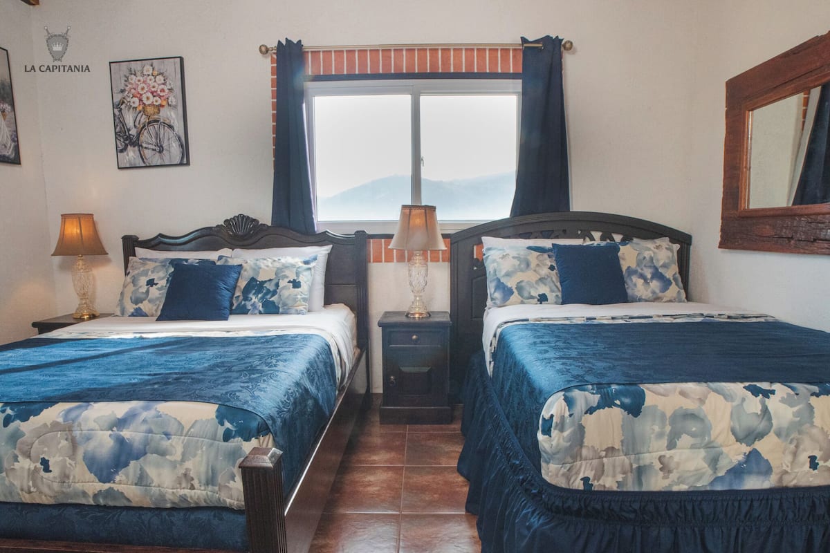A hotel room with two beds, a window with curtains, a lamp, and a wall with a framed picture.